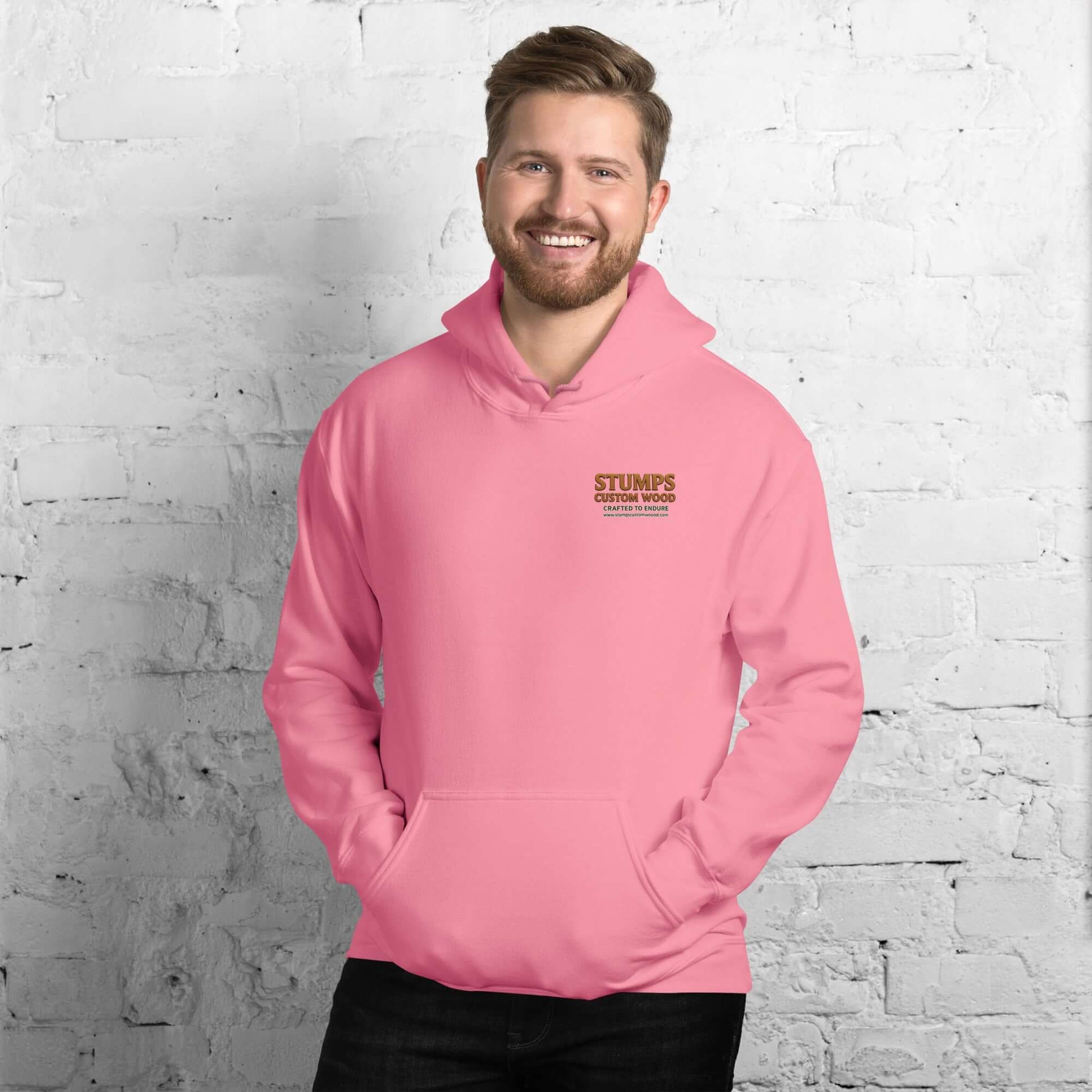 Stumps Custom Wood Unisex Hoodie in pink worn by a smiling man against a brick wall.